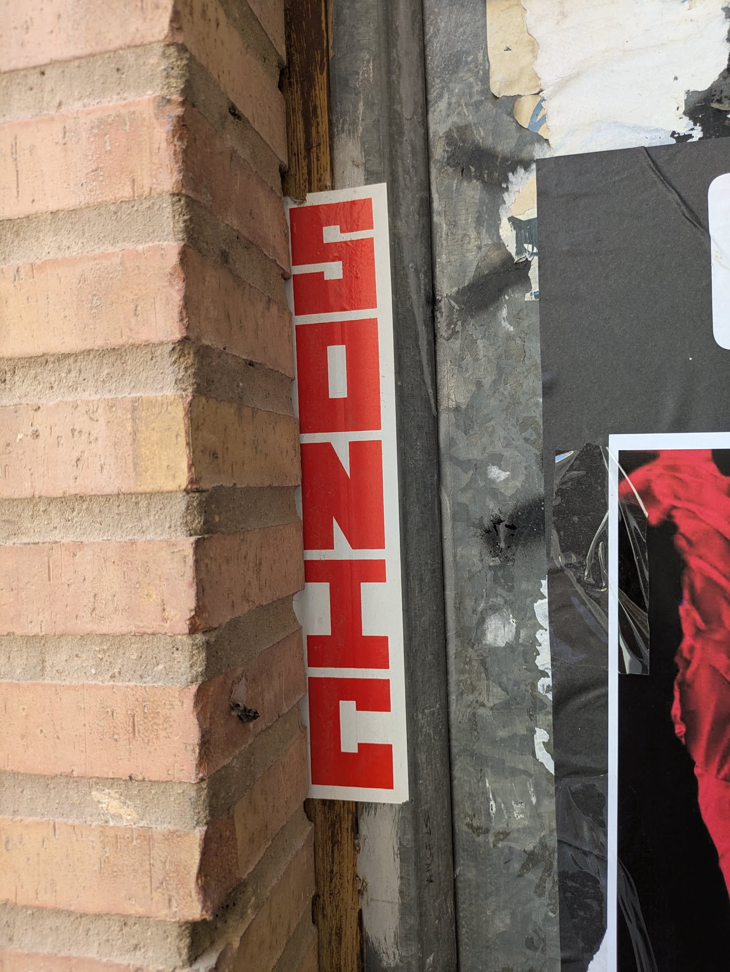 The sticker is red and white and vertically oriented, it spells the word 'SONHE' in Portuguese, meaning 'dream'. It's placed between the corner of a brick wall and a weathered metal surface with other posters.