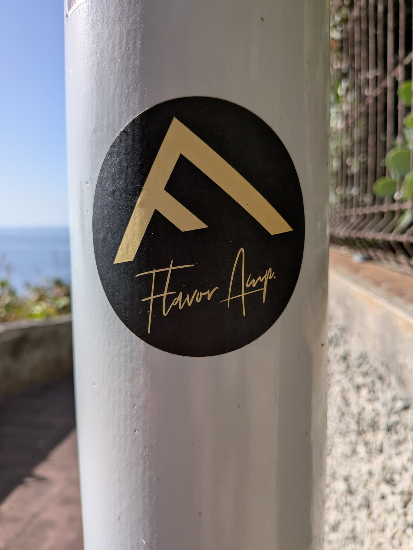 A circular black sticker affixed to a white post. It features a gold geometric logo resembling a stylized 'F' above the handwritten-style text 'Flavor Amp' in gold.