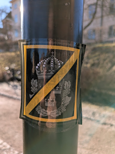 A black sticker with a gold border depicting the Swedish coat of arms, which includes a crown and crossed axes, is crossed out with a gold line. The sticker is affixed to a lamppost.