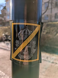 Street sticker A black sticker with a gold border depicting the Swedish coat of arms, which includes a crown and crossed axes, is crossed out with a gold line. The sticker is affixed to a lamppost.