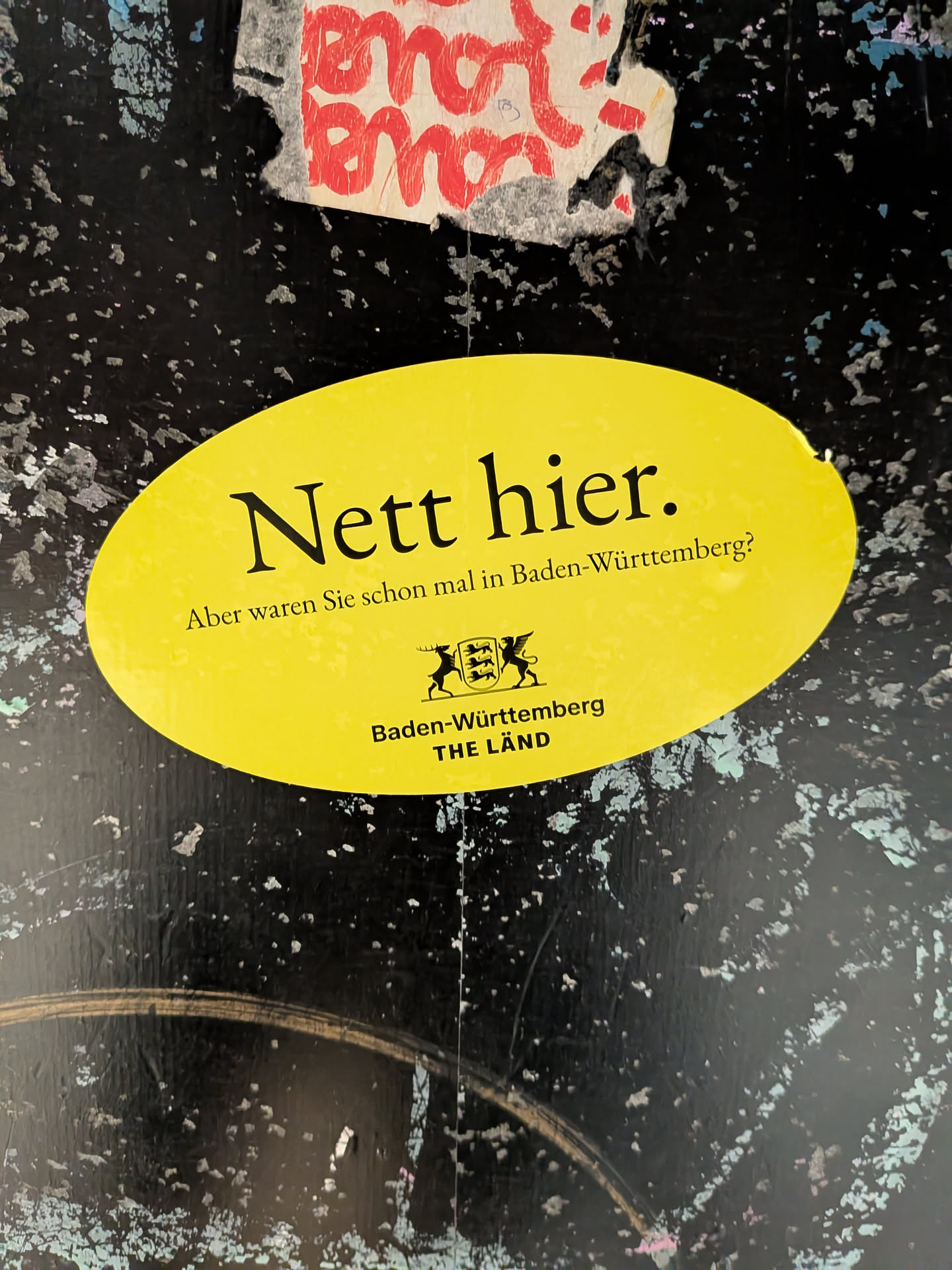 An oval yellow sticker with black text reading 'Nett hier. Aber waren Sie schon mal in Baden-Württemberg?'. Features the state's coat of arms and 'THE LÄND' branding at the bottom, stuck on a dark, weathered surface.