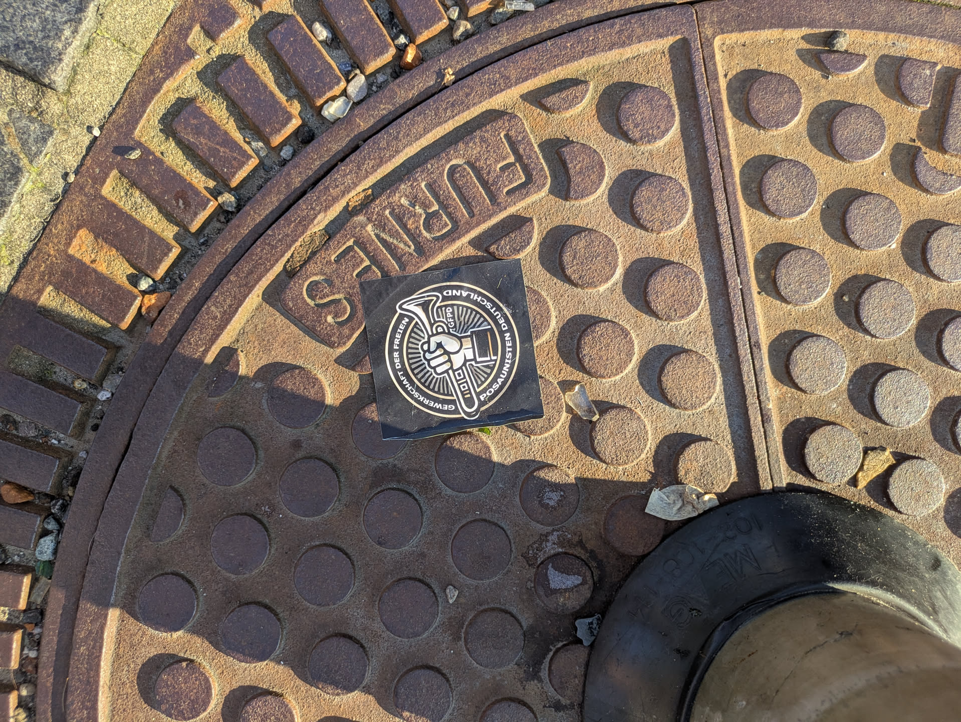 The image captures a square black sticker adhered to a textured, circular, rusty manhole cover. The sticker prominently displays a silver and white emblem: a stylized raised fist clutching what appears to be a trombone or a similar brass instrument, from which rays emanate. Around this central motif, in a circular arrangement, is text in German: 'GEWERKSCHAFT DER FREIEN POSAUNISTEN DEUTSCHLAND' (Union of Free Trombonists Germany). The initials 'GFFPD' are also visible within the emblem. The manhole cover itself has a pattern of raised circular bumps and an outer rim with rectangular segments. The word 'FURNES' is embossed on the manhole cover near the sticker. The lighting suggests sunlight, casting shadows that highlight the textures of both the sticker and the metal surface.