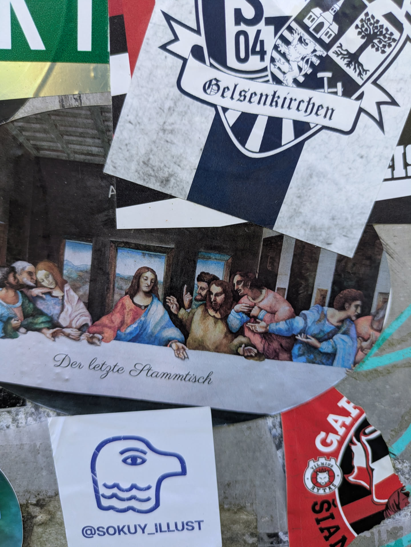 The central sticker displays a well-preserved parody of Leonardo da Vinci's renowned painting, 'The Last Supper.' The figures are depicted in poses and arrangements similar to the original masterpiece, seated at a long table. However, the scene is given a modern, humorous twist by the German text below, which reads 'Der letzte Stammtisch' (The last regulars' table or the last pub gathering). The artwork maintains a classic painting aesthetic with muted colors, contributing to its artistic feel. Below this main sticker, a smaller white sticker with a blue illustration of a head with a wave pattern is visible, and it features a social media handle.
