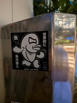 Street sticker Square black sticker with a white-outlined cartoon fish character in a dynamic pose. Features Japanese text on the sides and 'MERIOONE • FISHES INVASION' at the bottom.