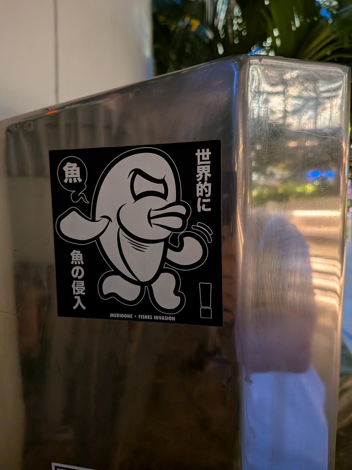 Square black sticker with a white-outlined cartoon fish character in a dynamic pose. Features Japanese text on the sides and 'MERIOONE • FISHES INVASION' at the bottom.