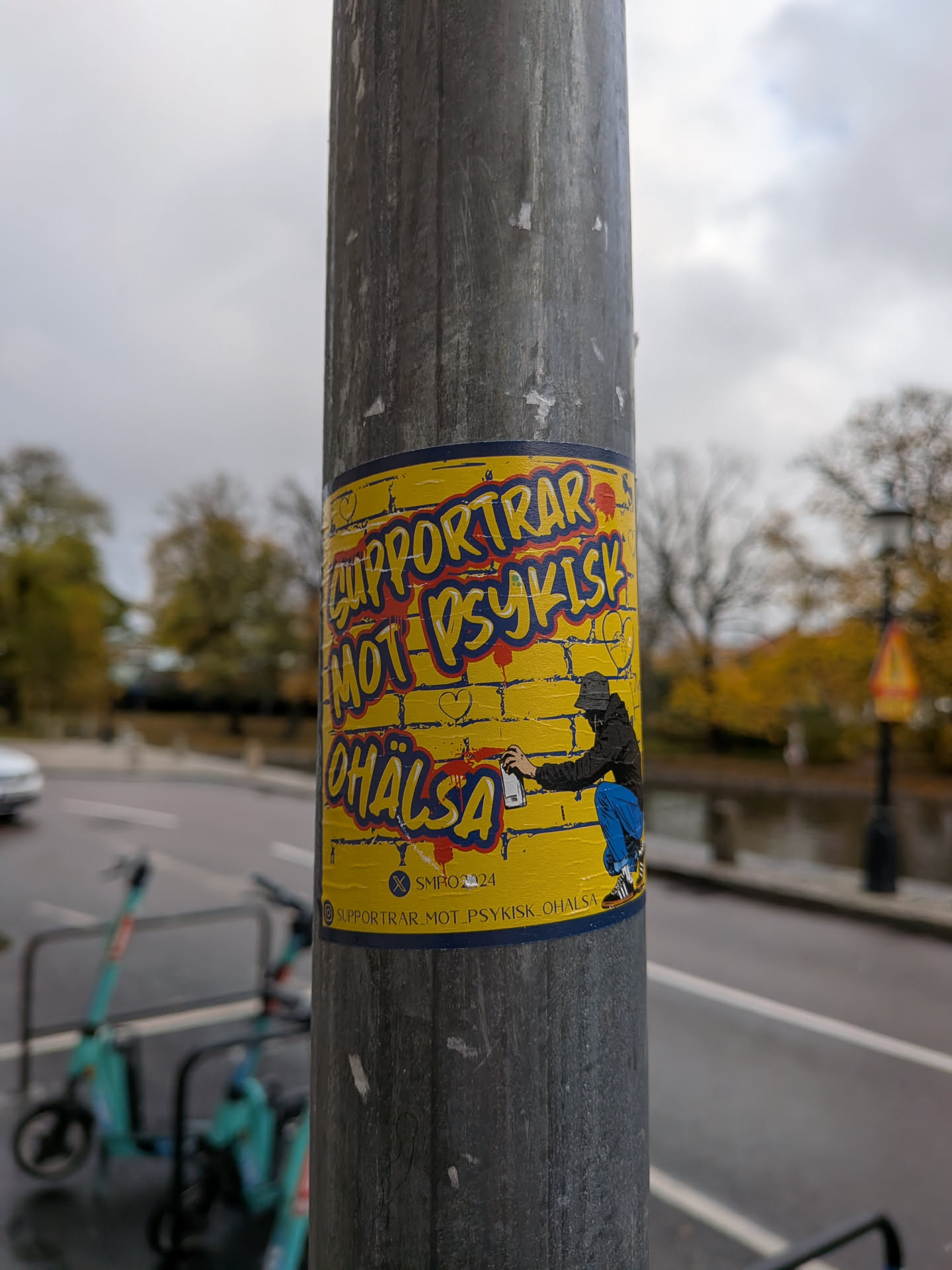 The sticker is predominantly yellow with a dark blue border. It features bold, stylized text in red and yellow with blue outlines that reads 'SUPPORTAR MOT PSYKISK OHÄLSA'. Below the main text, there's a graphic of a person wearing a black hoodie and blue pants, crouching and spray-painting. The background of the sticker appears to be a brick wall texture. There are also small red heart shapes scattered around the text. At the bottom of the sticker, there's a smaller line of text and a symbol.