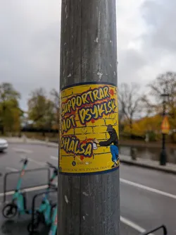 Street sticker The sticker is predominantly yellow with a dark blue border. It features bold, stylized text in red and yellow with blue outlines that reads 'SUPPORTAR MOT PSYKISK OHÄLSA'. Below the main text, there's a graphic of a person wearing a black hoodie and blue pants, crouching and spray-painting. The background of the sticker appears to be a brick wall texture. There are also small red heart shapes scattered around the text. At the bottom of the sticker, there's a smaller line of text and a symbol.
