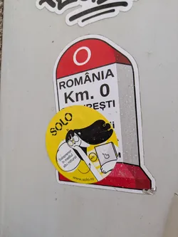 Street sticker A round, yellow sticker with a cartoon illustration of a woman holding a laptop. The sticker also has some text. The stickers is placed on another sticker representing the Kilometer Zero monument in Romania.
