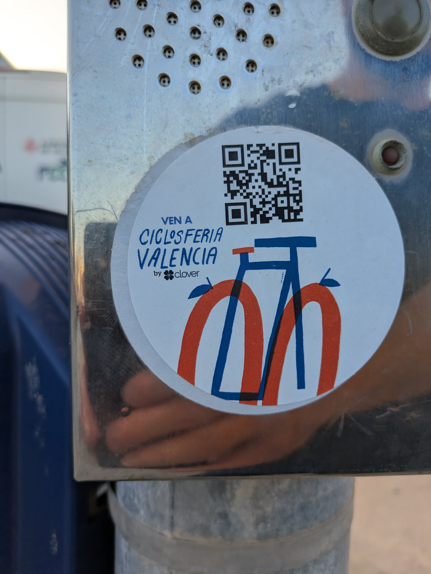A round sticker adhered to a metal surface advertises the Ciclosferia Valencia. The sticker features a QR code, the text "VEN A CICLOSFERIA VALENCIA by clover", and a stylized blue and red bicycle drawing. The bike is an artistic representation with curved lines for the wheels and a minimalist frame. The sticker is circular, with a white background.