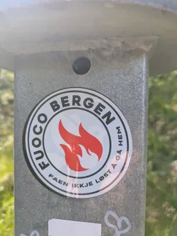 Street sticker A circular sticker with a white background and a bold black outer ring. The center features two stylized, vibrant red flames. Arching above the flames, the text "FUOCO BERGEN" is written in black capital letters. Below the flames, curved text in black capital letters reads "FAEN IKKJE LØST Å GÅ HEM". The sticker is affixed to a textured grey metal pole.
