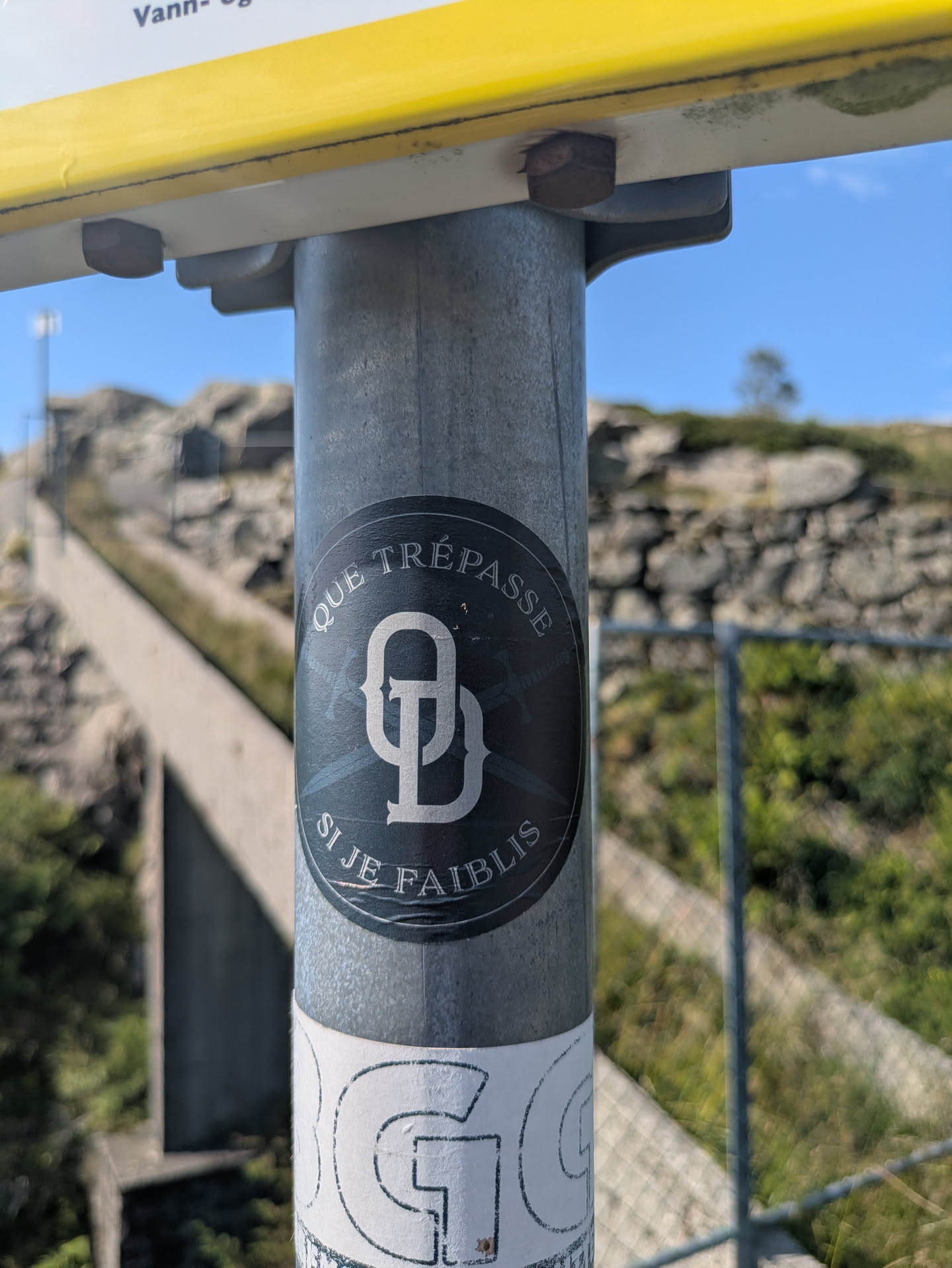 The sticker is dark in color, possibly black or dark grey, with light grey or white text and a prominent monogram in the center. The text, arranged in an arc at the top and bottom, reads "QUE TRÉPASSE SI JE FAIBLIS". Behind the stylized monogram, which appears to be 'GD' or 'QD', there are subtle cross-hatched lines or possibly faint crossed swords. The sticker is affixed to a textured grey metal pole, with a blurred outdoor background showing a path, rocks, and blue sky.