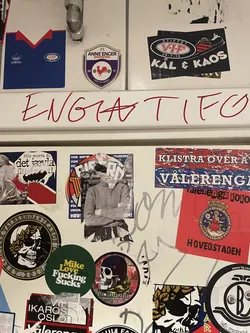 Street sticker The central sticker features a person, likely male, depicted from the chest up, wearing a plain grey turtleneck sweater. Their arms are crossed over their chest. The most prominent detail is the person's face, which is largely obscured and appears to be torn or collaged with white paper fragments, creating a distressed and abstract visual. The background immediately behind the figure is dark and indistinct, suggesting an urban or textured surface. The edges of the sticker itself show signs of wear and tear, indicating it's a physical sticker that has been applied and possibly partially damaged over time.