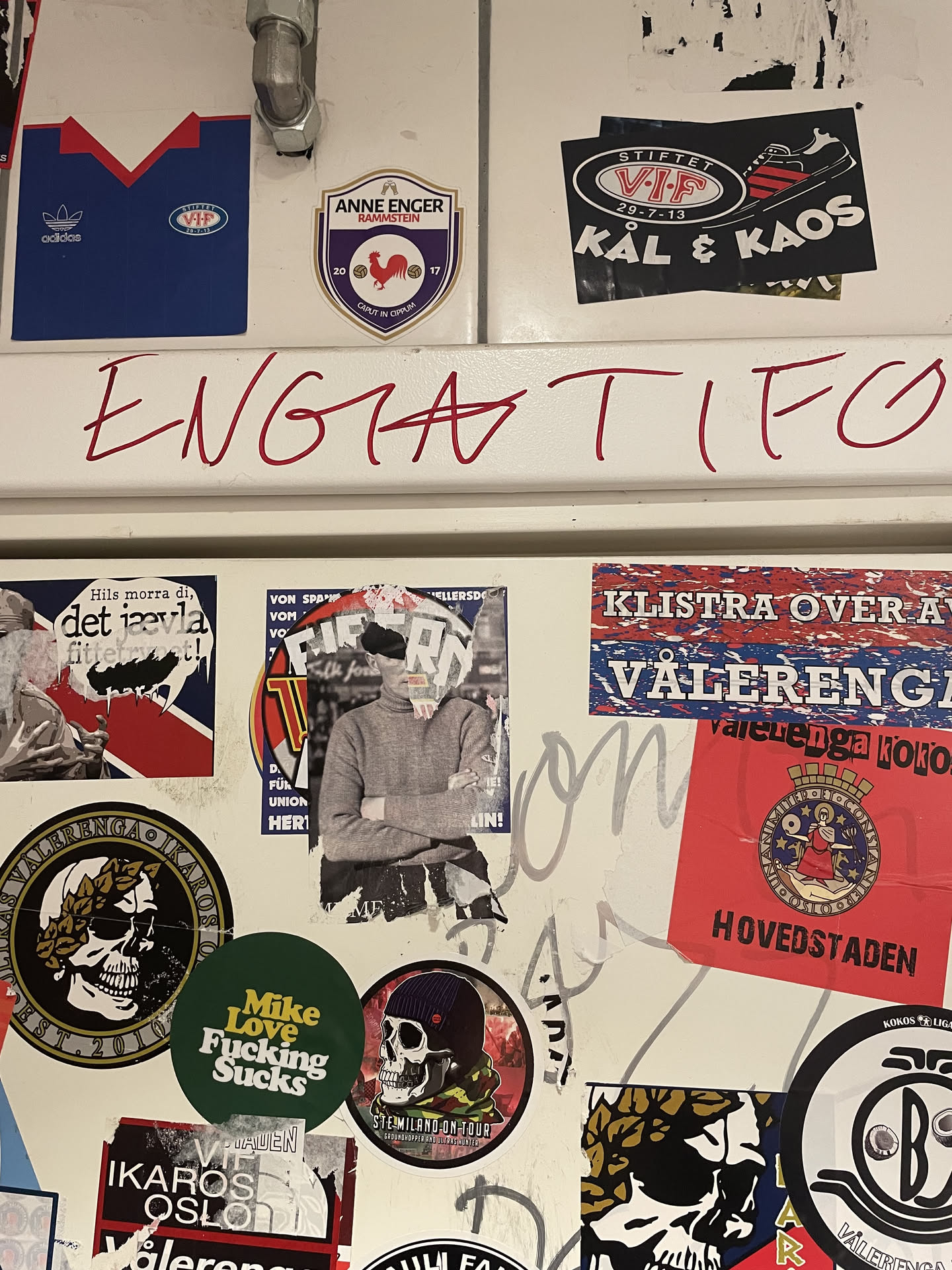 The central sticker features a person, likely male, depicted from the chest up, wearing a plain grey turtleneck sweater. Their arms are crossed over their chest. The most prominent detail is the person's face, which is largely obscured and appears to be torn or collaged with white paper fragments, creating a distressed and abstract visual. The background immediately behind the figure is dark and indistinct, suggesting an urban or textured surface. The edges of the sticker itself show signs of wear and tear, indicating it's a physical sticker that has been applied and possibly partially damaged over time.