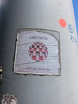 Street sticker A square, wrinkled paper sticker on a grey pole. It displays a central pink and black checkered sphere, with 'AMETROM' written above and 'ATELIER ELEKTRO' below.