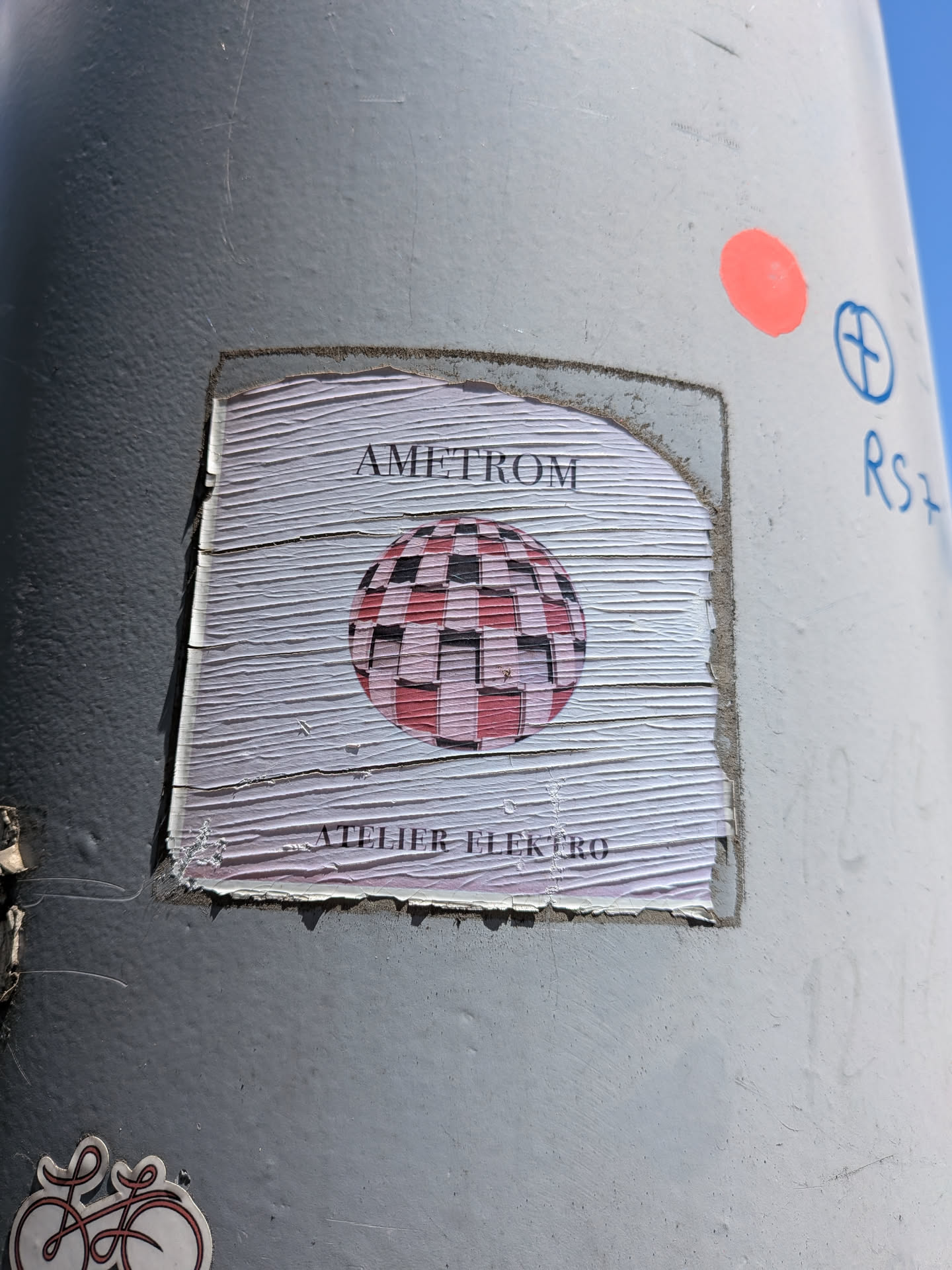 A square, wrinkled paper sticker on a grey pole. It displays a central pink and black checkered sphere, with 'AMETROM' written above and 'ATELIER ELEKTRO' below.