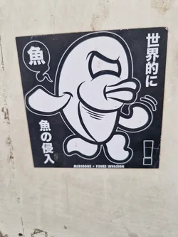A black and white sticker featuring a cartoon fish with an angry expression. The text on the sticker is in Japanese and translates to "World domination, fish invasion". The artist's name or brand is MERIOONE.