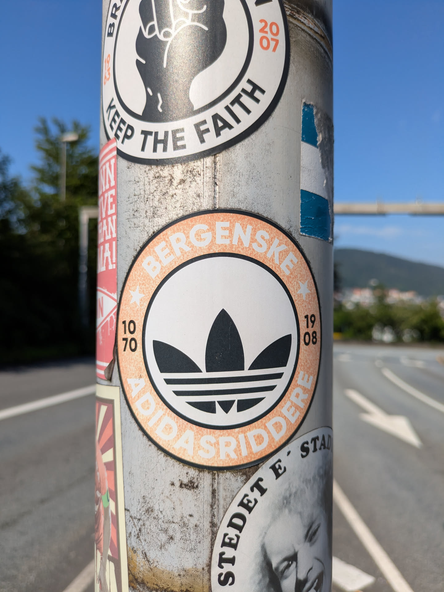 The central sticker is circular, with a distinctive Adidas Trefoil logo in black on a white background. It has an orange-brown outer ring with white text that reads "BERGENSKE" on the top arc and "ADIDASRIDDERE" on the bottom arc. To the left of the logo, the numbers "10 70" are vertically aligned, and to the right, "19 08" are similarly aligned. The sticker appears worn, showing some texture and light damage, indicative of being outdoors on a utility pole. The pole itself is metallic and bears other, non-focused stickers.