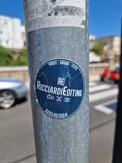 Street sticker A blue round sticker with the text RicciardieEditing, Video - Drone - Foto, and the number 3201421504. It features icons for a camera, a drone, and a video camera. The sticker is affixed to a metal pole.