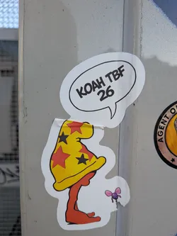 Street sticker The sticker features a stylized, abstract character with an orange-red, blob-like body that appears to be sitting or crouching. It wears a prominent, pointed yellow hat adorned with a pattern of red and black stars. The hat has a wide brim. Above the character's head, a white speech bubble with a black outline contains text. To the right of the character's head, a small, purple, insect-like creature with pink bow-like wings is depicted. The sticker has a white border and shows signs of wear, with some edges slightly peeled, indicating it's a physical sticker on a pole. The overall style is cartoonish and playful.