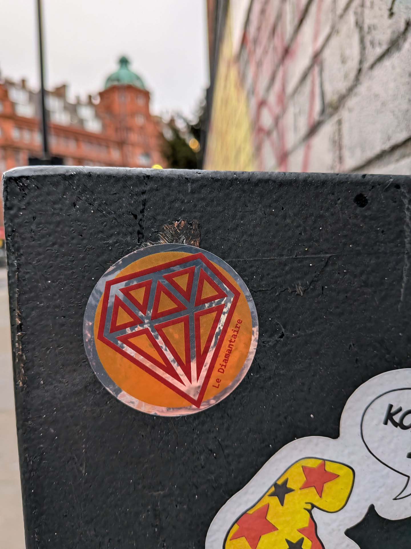 The sticker is circular, featuring a prominent, stylized diamond graphic at its center. The diamond is rendered with a silver-grey outline and internal facets, filled with a vibrant orange color. Some facets are also outlined in red, creating a layered, geometric effect. The background of the circular sticker is a slightly darker shade of orange. The silver-grey elements of the diamond appear reflective. Along the right edge of the circle, oriented vertically, is the text 'Le Diamantaire'. The sticker shows some signs of wear and is adhered to a textured, dark surface.