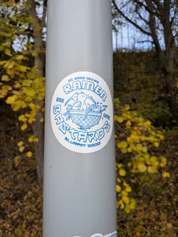 Street sticker The sticker is circular with a white background and blue lettering and illustrations. The central image depicts two cartoon birds dipping their heads into a bowl of ramen noodles. The bowl has a traditional Japanese wave pattern. Around the central image, text is arranged in a circular fashion. The top arc reads 'DC 2002 RECIPE RAMEN'. The bottom arc reads 'SLURPIN' GOOD!'. To the left of the bowl, the number '696' is written vertically. To the right of the bowl, the word 'UME' is written vertically. Below the bowl, in a larger font, are the words 'BASTARDS' in a stylized, blocky font.