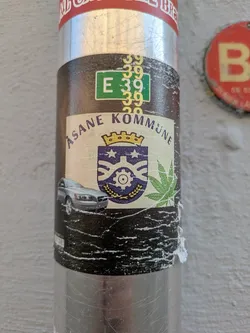 Street sticker The sticker, affixed to a metal pole, features a central cream-colored banner with the text "ÅSANE KOMMUNE" curved above a blue and gold crest or coat of arms, topped with a crown. To the left of the crest is a silver Volvo car, and to the right is a prominent green cannabis leaf. Above the banner, a green rectangle displays "E 39", surrounded by several instances of the number "39" in yellow. The background of the sticker below the banner is dark brown or black, showing signs of wear and peeling.