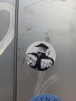 The sticker depicts a figure, possibly wearing a military or formal cap, with their eyes obscured by shadow or a visor. Their hands are clenched into fists, prominently displaying the words 'SAME' tattooed across the knuckles of the left hand and 'PAIN' across the knuckles of the right hand. The image is rendered in black and white, giving it a stark and impactful appearance. It is placed on a brushed metal surface.