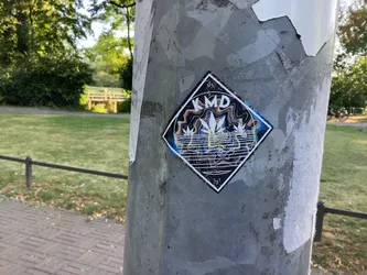 Street sticker The sticker is square with its corners pointing up and down, creating a diamond shape. It features a white cannabis leaf in the center, surrounded by swirling, psychedelic patterns in shades of blue, yellow, and brown. The word 'KMD' is prominently displayed in white capital letters above the leaf. There is also text '383' above the 'KMD' and '581' below the leaf, all in small white font. The sticker has a white border and appears to be made of paper.