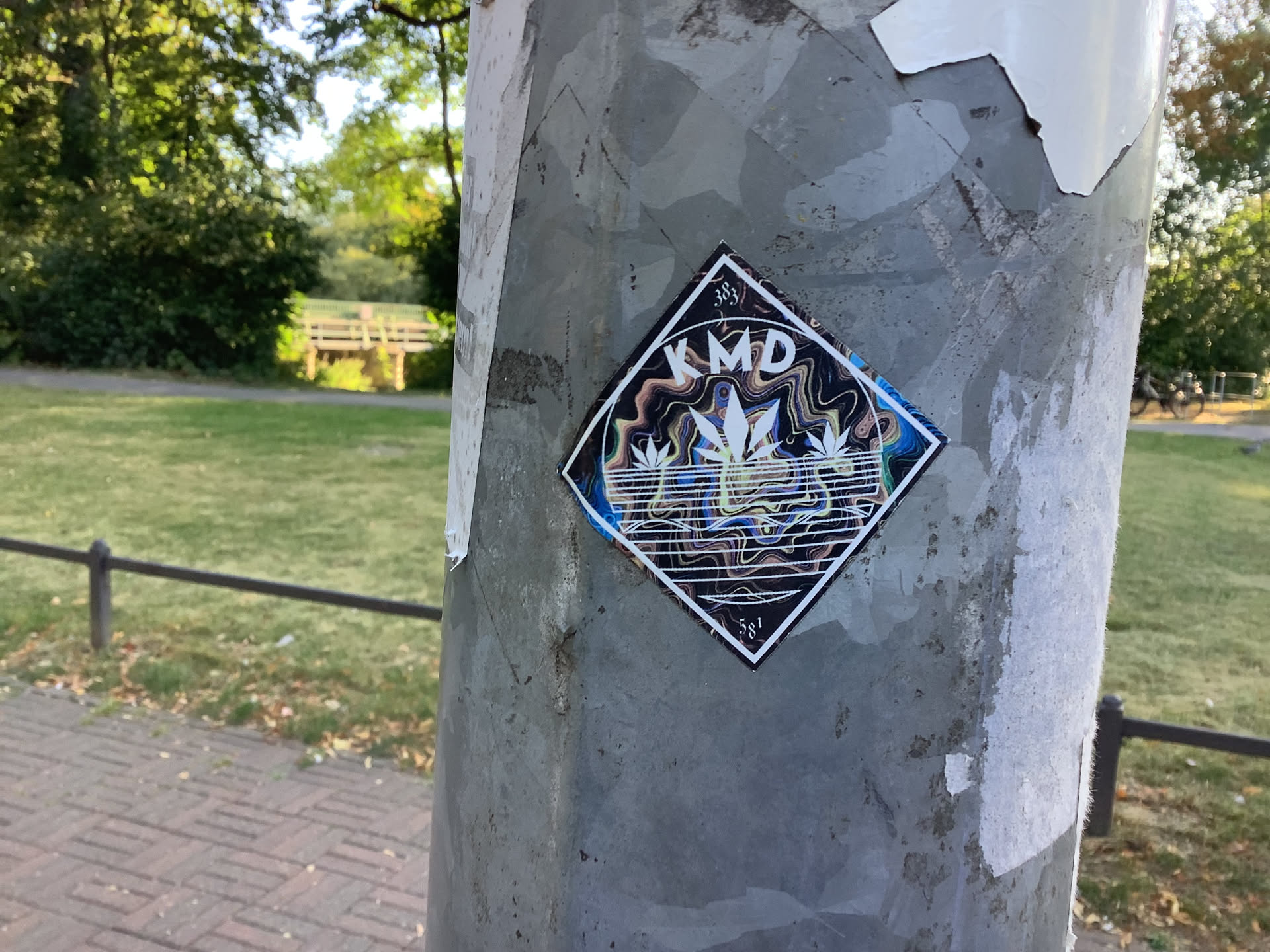 The sticker is square with its corners pointing up and down, creating a diamond shape. It features a white cannabis leaf in the center, surrounded by swirling, psychedelic patterns in shades of blue, yellow, and brown. The word 'KMD' is prominently displayed in white capital letters above the leaf. There is also text '383' above the 'KMD' and '581' below the leaf, all in small white font. The sticker has a white border and appears to be made of paper.