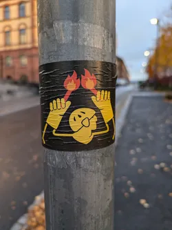 A black and yellow sticker depicting a simple cartoon character holding two flaming torches. The sticker is affixed to a weathered grey metal pole.  The style is minimalist and playful. The background shows an out-of-focus street scene.