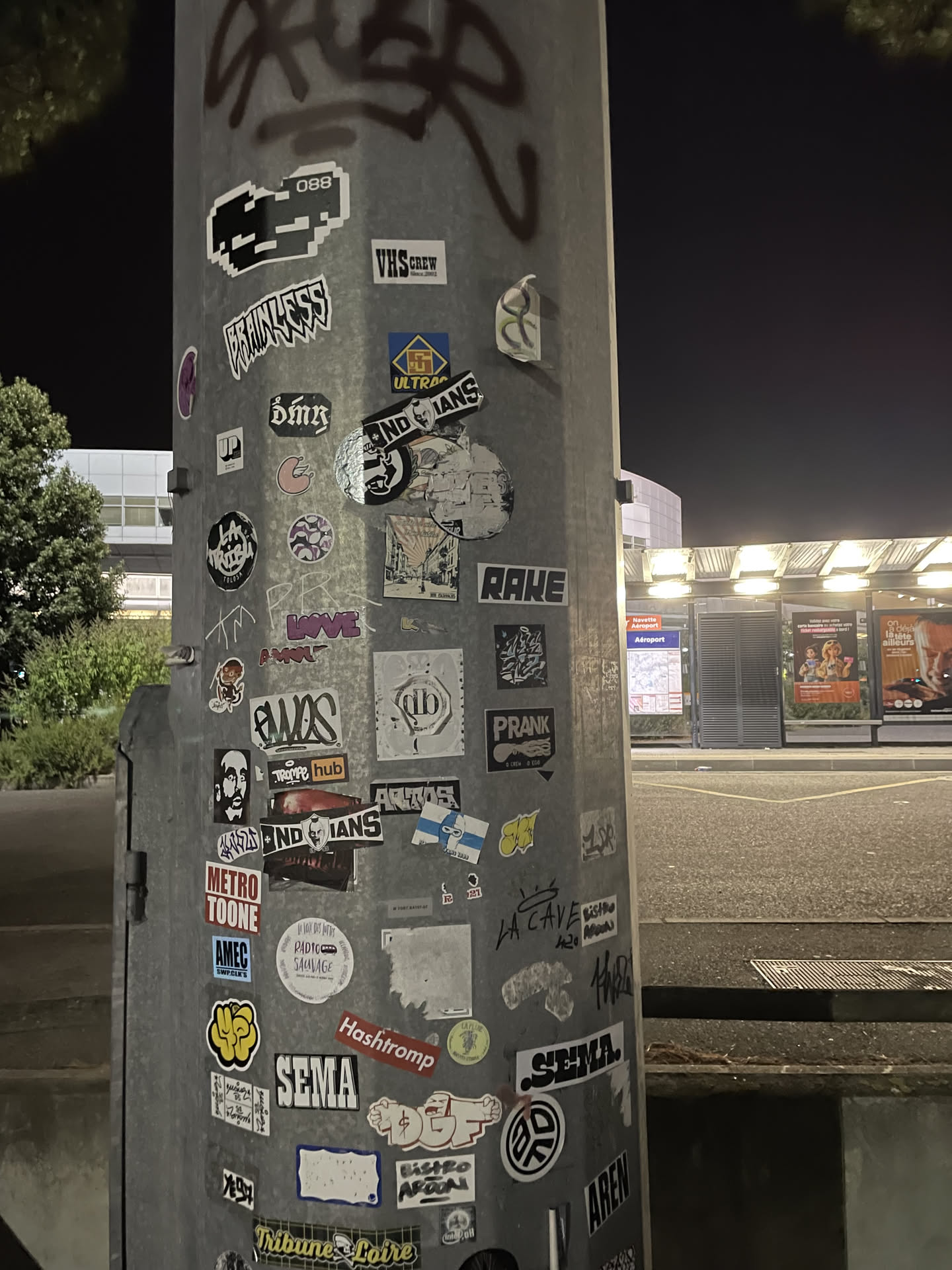 This circular sticker is rendered in stark black and white, depicting a highly stylized, almost abstract human face. The face features prominent, dark eyes and is adorned with what appears to be a feathered headdress or an elaborate hair arrangement, giving it an indigenous or tribal aesthetic. The word "NDIANS" is integrated into the lower part of the design, written in a strong, sans-serif font. The edges of the sticker show some minor wear and tear, and it is partially obscured by other stickers and graffiti on the pole.