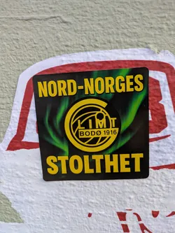 Street sticker The sticker is rectangular, predominantly black with a vibrant green swirling pattern behind a central yellow circular logo. The logo contains the text "GLIMT" and "BODØ 1916". Above the logo, in large yellow capital letters, is "NORD-NORGES", and below it, also in large yellow capitals, is "STOLTHET". The sticker is applied to a light-colored, textured wall and partially overlaps a faded red and white sticker underneath.