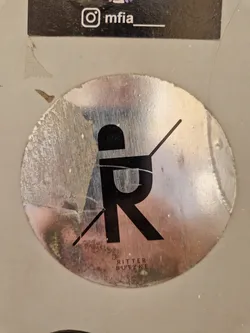 A round, silver sticker with a minimalist black logo featuring the letters 'R' and 'B' intersecting.  The text 'Ritter Butzke' is visible below the logo. The sticker shows some signs of wear and tear, adding to its unique character. An Instagram handle (@mfia) is visible on a sticker above the main sticker.