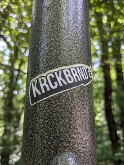 A dark gray and white sticker with the text 'KRCKBRND' is affixed to a textured gray pole in a forest setting. The sticker has a slightly curved rectangular shape with a small symbol near the end of the text. The background is blurred and shows the green foliage of the trees.
