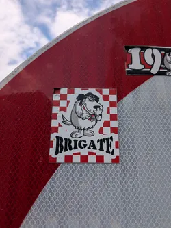 Street sticker Square sticker with a red and white checkered background. Features the cartoon dog Muttley wearing aviator goggles and snickering. The word "BRIGATE" is printed below in bold black font.