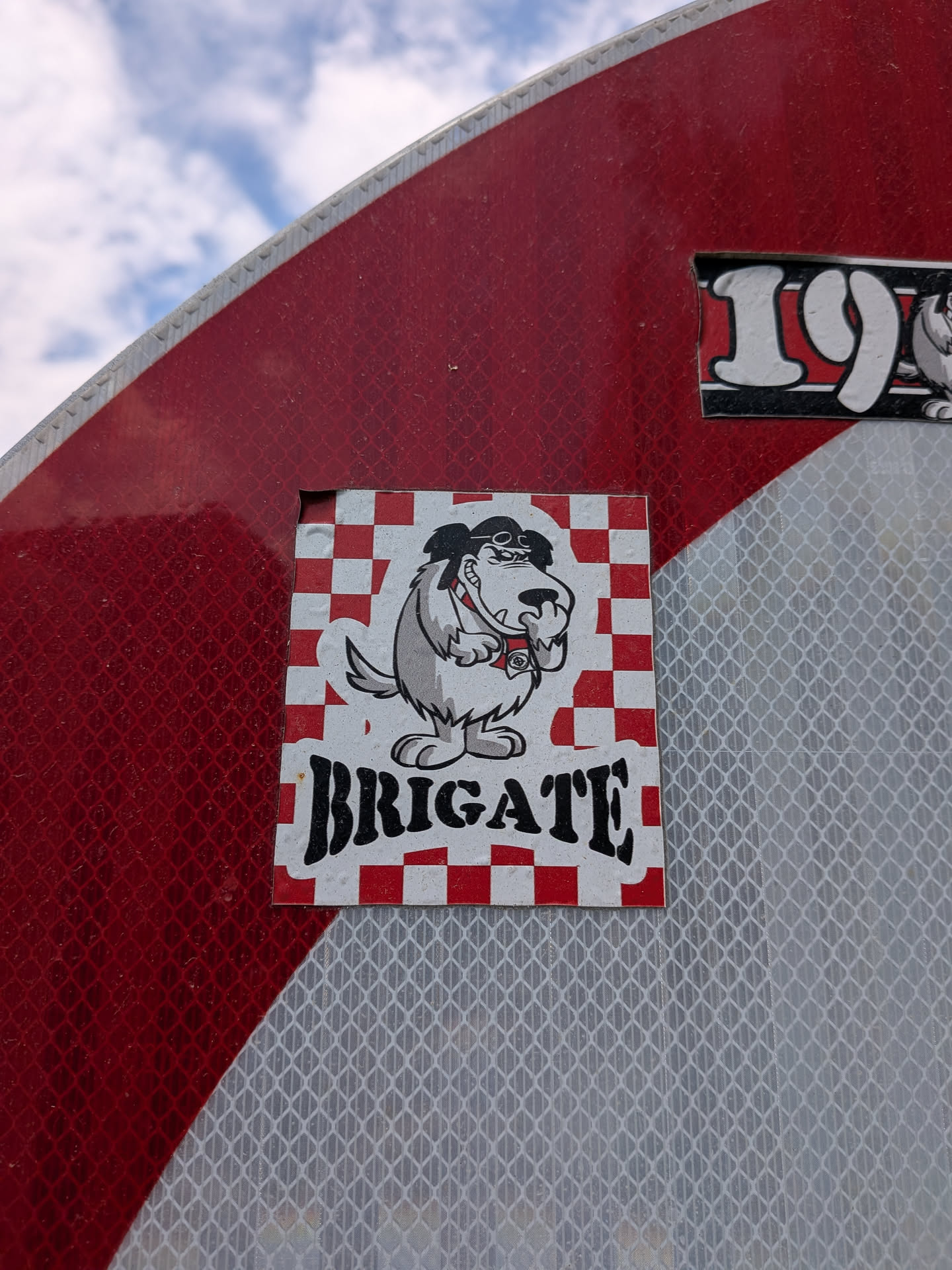 Square sticker with a red and white checkered background. Features the cartoon dog Muttley wearing aviator goggles and snickering. The word "BRIGATE" is printed below in bold black font.
