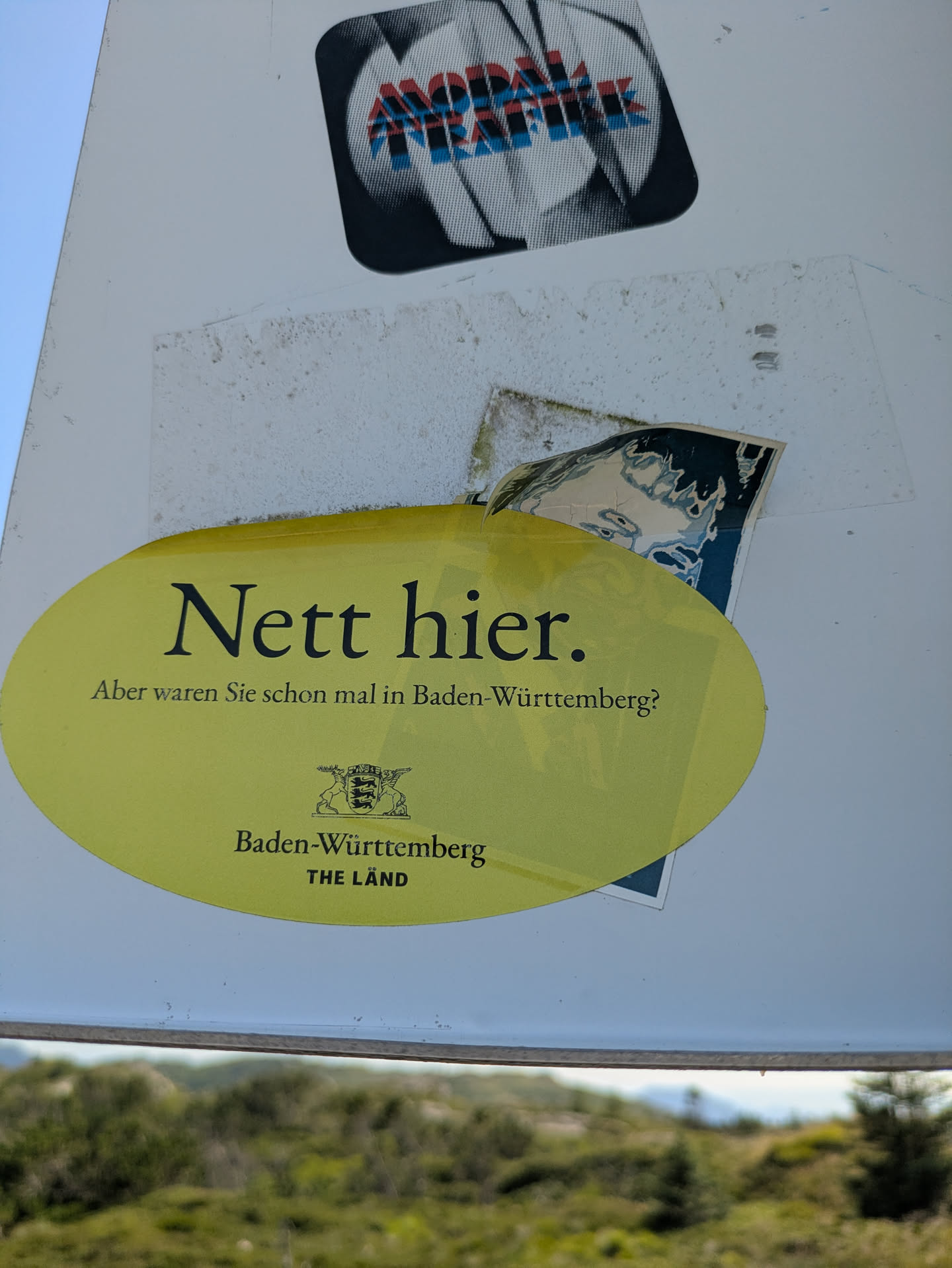 The sticker prominently featured in the center of the image is an oval-shaped, bright yellow decal. It displays black text in varying sizes. The largest text reads "Nett hier." followed by smaller text asking, "Aber waren Sie schon mal in Baden-Württemberg?". Below this, the coat of arms of Baden-Württemberg (three lions) is depicted, beneath which the words "Baden-Württemberg" and "THE LÄND" are printed in smaller black capital letters. The sticker appears slightly worn and is affixed to a white surface, possibly a pole or sign, set against a blurred background of green foliage under a clear sky.