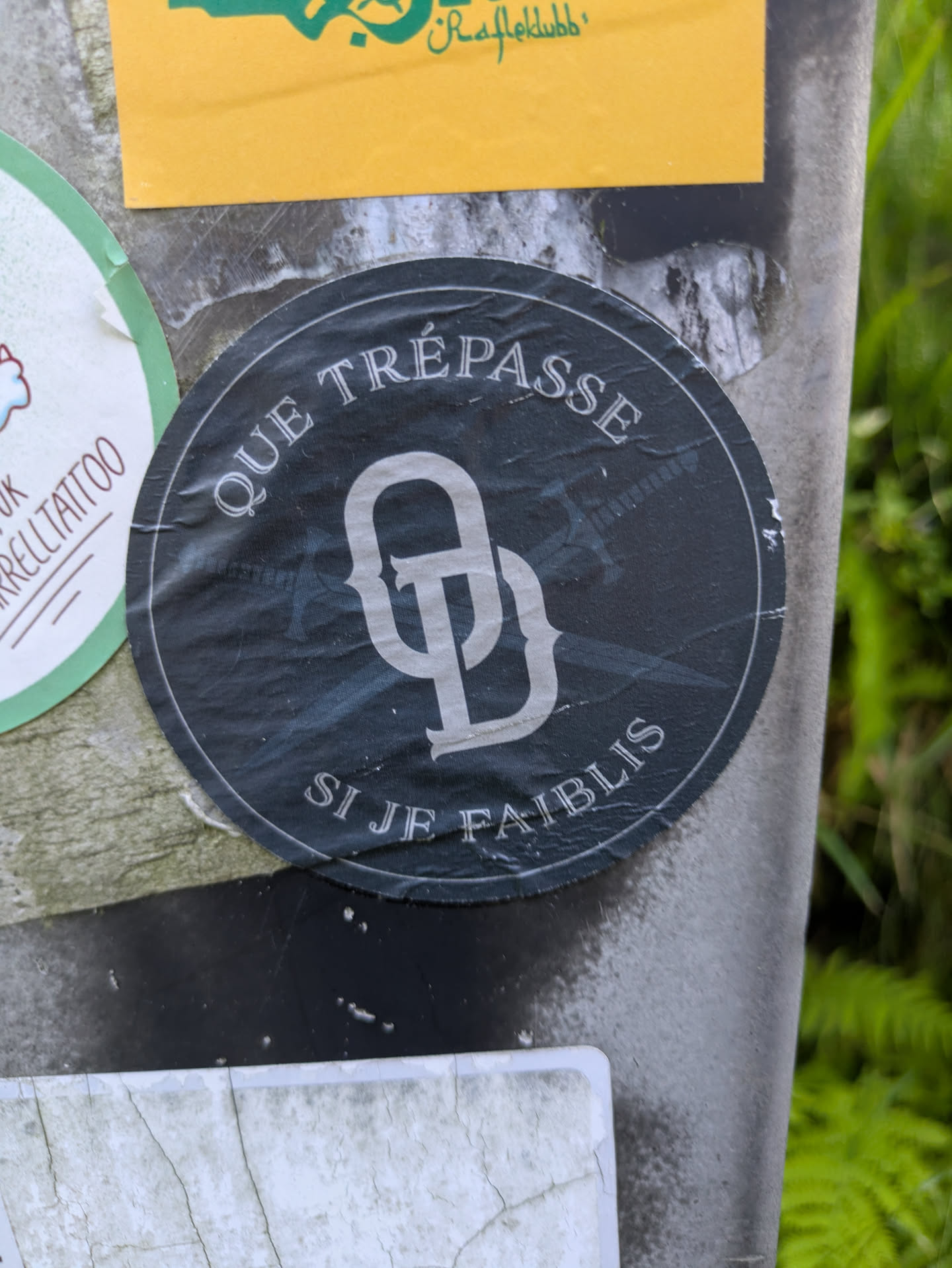 The sticker is round with a dark grey or black background and white text. The phrase 'QUE TRÉPASSE' is arched across the top, and 'SI JE FAIBLIS' is arched across the bottom. In the center, there is a prominent, stylized white monogram, appearing to be a decorative combination of the letters 'Q' and 'D' or 'P'. The sticker is slightly creased and shows signs of wear.