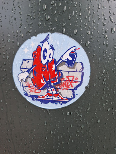 Round sticker featuring a cartoon character in the style of 80s and 90s graffiti art. The character is red and white, with big eyes and is holding a spray can. The background is a light blue with a graffiti wall behind the character. The sticker is attached to a dark-colored surface with water droplets on it.