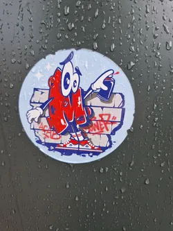 Street sticker Round sticker featuring a cartoon character in the style of 80s and 90s graffiti art. The character is red and white, with big eyes and is holding a spray can. The background is a light blue with a graffiti wall behind the character. The sticker is attached to a dark-colored surface with water droplets on it.