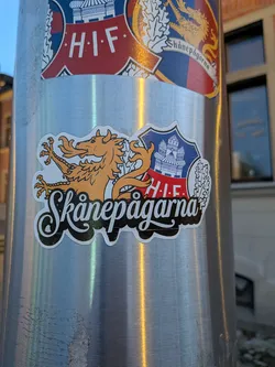 Street sticker The sticker features a stylized golden dragon with sharp claws and a long, serpentine body, positioned to the left of a shield. The shield is divided into sections: the top portion is blue with a white depiction of a castle or fortress, and the bottom portion is red with white text 'H.I.F.'. To the right of the shield is a white, stylized oak leaf. Below the dragon and shield, the text 'Skåne pågarna' is written in a decorative, white script with a black outline. The entire sticker has a white border.