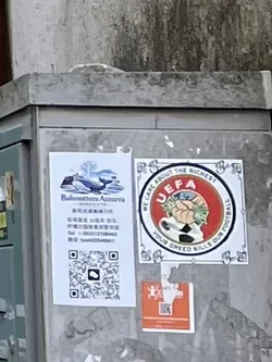 The central sticker is square with a decorative white border featuring blue floral or scroll-like patterns in each corner and along the edges. Inside this border, there is a circular emblem with a vibrant red background. This emblem parodies the UEFA logo, depicting a figure whose hands are clutching large wads of green cash, emerging from what appears to be a stylized football or globe. The letters 'UEFA' are prominently displayed in white at the top of the red circle. Arcing around the red circle, within the white border, is text that reads 'WE CARE ABOUT THE RICHEST' at the top and 'YOUR GREED KILLS OUR FOOTBALL' at the bottom. The overall color palette includes red, white, blue, and green.