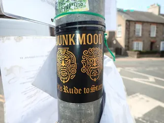 Street sticker A black vinyl sticker wrapped around a metal post. It displays the text 'JUNKMOOD' in gold at the top, two gold-outlined tribal mask illustrations in the center, and the phrase 'It's Rude to Stare' in a gothic font at the bottom.