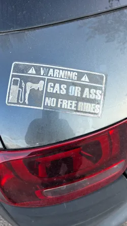 Street sticker A rectangular, weathered sticker affixed to a grey car body above a red taillight. The sticker is designed to look like a warning label, with a dark background and white text/graphics. At the top, it reads 'WARNING' flanked by two warning triangle icons. Below, on the left, is a graphic depicting a gas pump and a silhouette of a person bent over. On the right, the text reads 'GAS OR ASS' on the first line and 'NO FREE RIDES' on the second line. The sticker shows signs of aging and dirt.