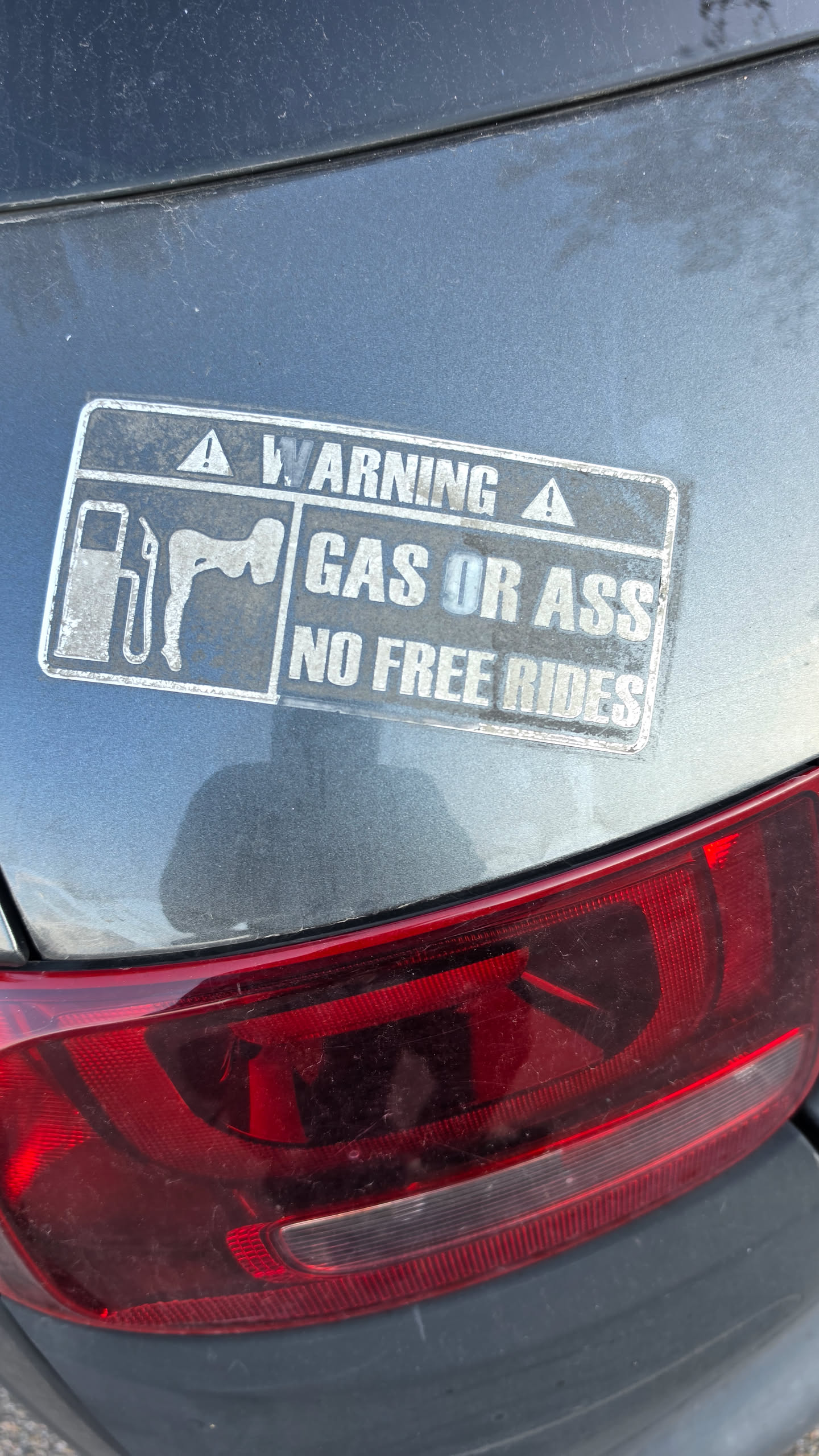 A rectangular, weathered sticker affixed to a grey car body above a red taillight. The sticker is designed to look like a warning label, with a dark background and white text/graphics. At the top, it reads 'WARNING' flanked by two warning triangle icons. Below, on the left, is a graphic depicting a gas pump and a silhouette of a person bent over. On the right, the text reads 'GAS OR ASS' on the first line and 'NO FREE RIDES' on the second line. The sticker shows signs of aging and dirt.