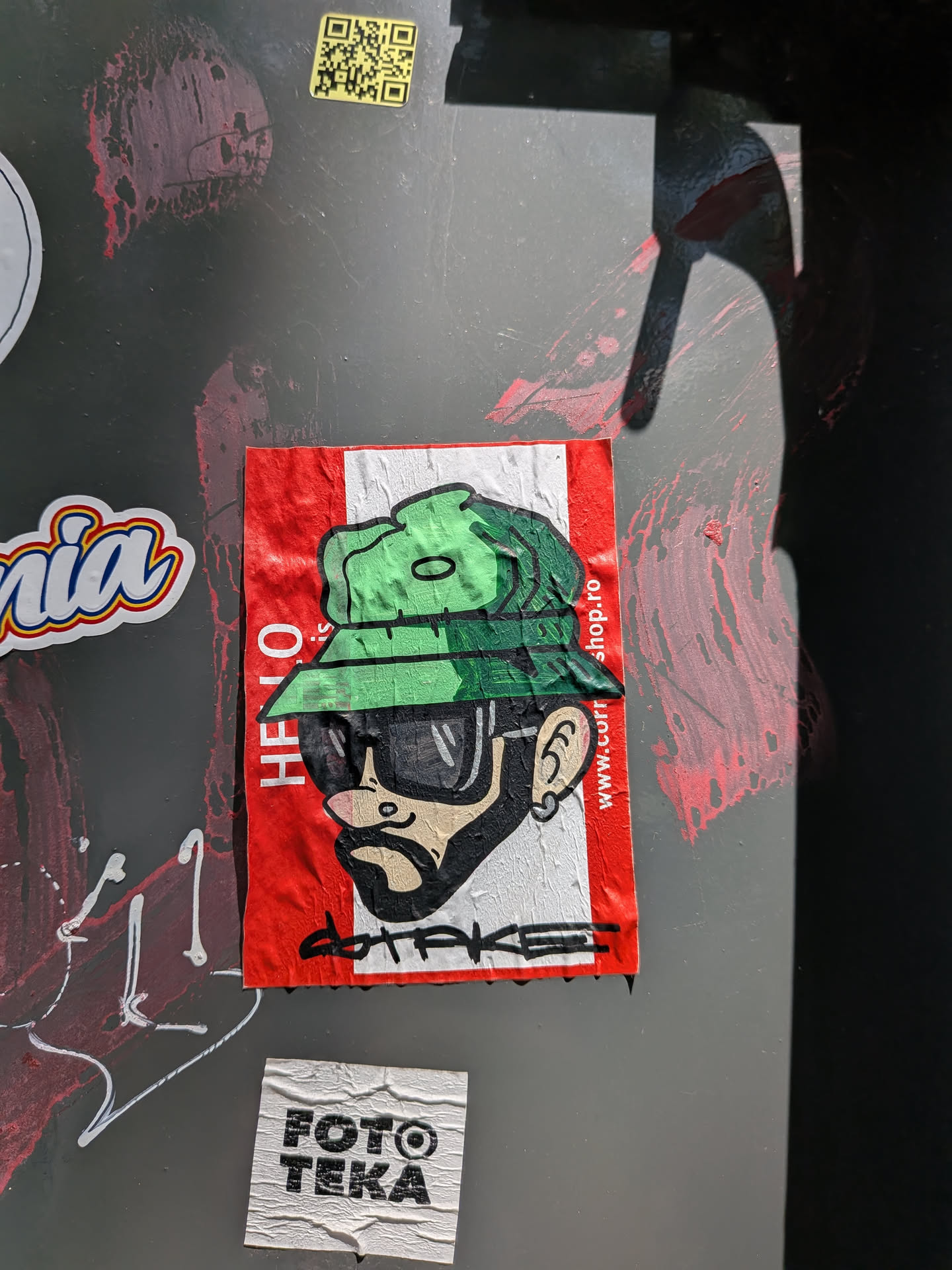 A rectangular sticker with a red and white background, styled like a name badge. It depicts a bearded character wearing a green bucket hat and dark sunglasses. Vertical text runs along both sides, and a black graffiti signature is at the bottom.