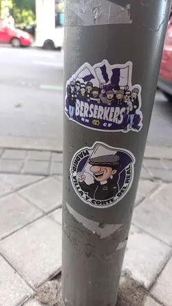 Street sticker The image showcases two distinct, cartoon-style stickers adhered to a weathered grey pole. The upper sticker is rectangular with rounded corners, depicting a crowd of stylized football supporters, or 'ultras', in various poses, some holding white and purple flags. They are gathered behind a banner that prominently displays 'BERSERKERS' in white capital letters, with 'RM' (featuring a crown logo) and 'CF' beneath it. The characters wear a mix of checkered caps, beanies, and sunglasses, rendered in purple, black, white, and skin tones. The lower sticker is circular, featuring a cheerful cartoon man with a large red nose, wearing a checkered flat cap and a dark jacket, holding a white flag with a purple emblem. Around him, in a circular arrangement, is the text 'MADRID, VILLA Y CORTE DEL REAL'. Both stickers are outlined in white, giving them a clean, die-cut appearance against the textured pole.