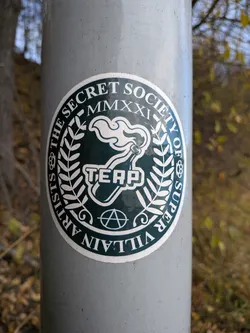 Street sticker The sticker is circular with a dark green background and white lettering and graphics. The outer ring of the sticker contains the text 'THE SECRET SOCIETY OF SUPER VILLAIN ARTISTS' in white, capital letters, arranged in a curve. Inside this ring, on the left and right sides, are decorative elements resembling laurel branches. At the top of the inner circle, 'MMXXI' is written in Roman numerals. The central graphic features a stylized white torch with flames, and below it, the word 'TEAP' in white, block letters. At the very bottom of the inner circle, an anarchy symbol (a circle with an 'A' inside) is depicted in white. The overall design suggests a clandestine or rebellious artistic collective.
