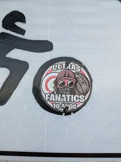 Street sticker A round, slightly worn sticker featuring a central character wearing a helmet, goggles, and face mask, positioned next to a football. The background consists of red and white concentric circles, framed by a black border. Text at the top reads 'ULTRAS', while the bottom reads 'FANATICS 10 ANOS'.