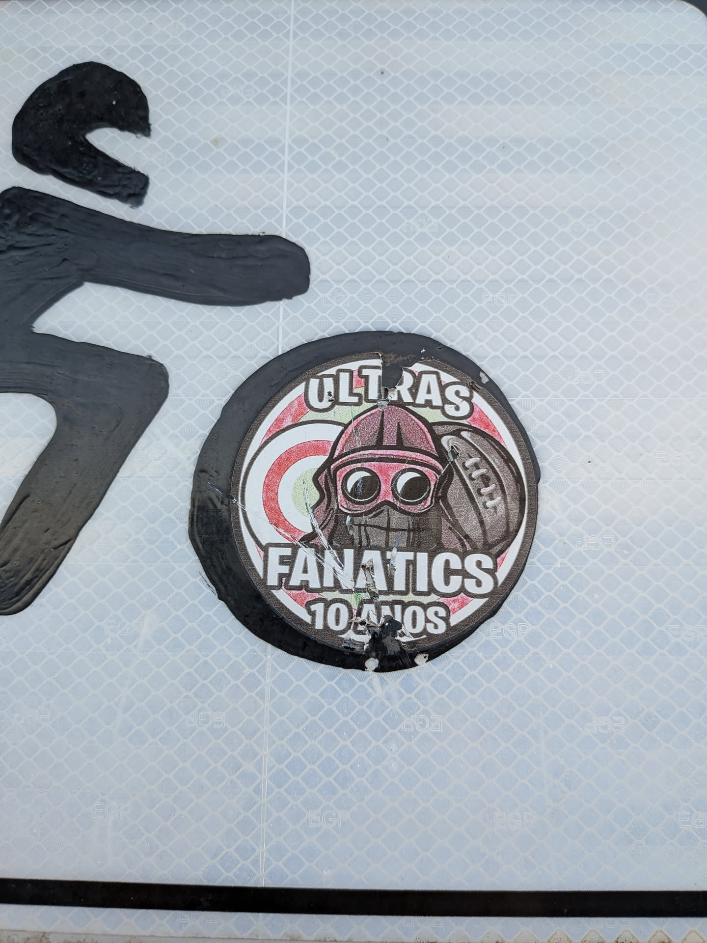 A round, slightly worn sticker featuring a central character wearing a helmet, goggles, and face mask, positioned next to a football. The background consists of red and white concentric circles, framed by a black border. Text at the top reads 'ULTRAS', while the bottom reads 'FANATICS 10 ANOS'.